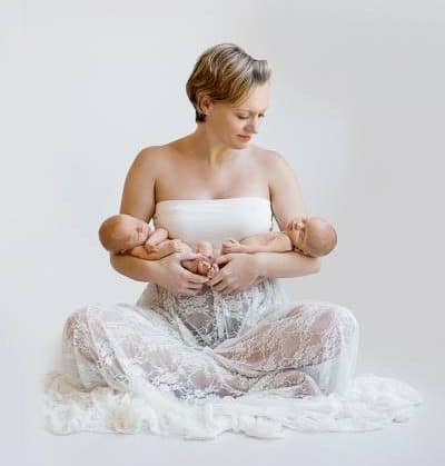A mother sitting cross-legged in a beautiful white gown, holding her newborn twins.