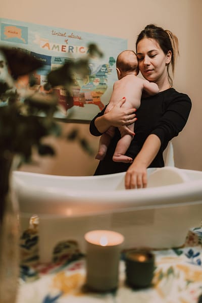 A young mother preparing a warm bath, while she holds her baby, surrounded by candles