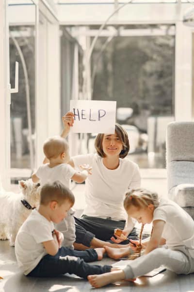 A humerous image of a mother holding up a "help" sign, sitting on the living floor with her 3 young children.