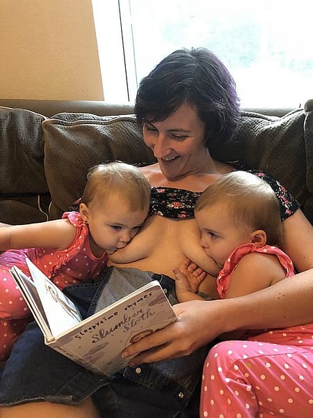 A mother, tandem feeding twins, while sitting on the couch, reading the babies a story