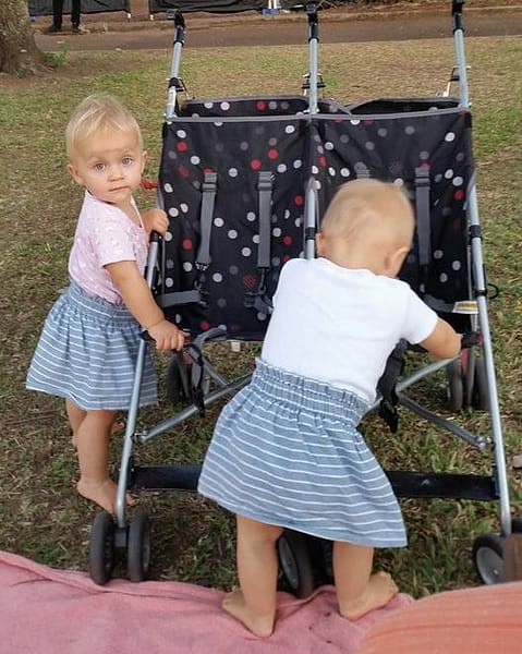Cute twin toddlers, holding on to a twin pram, as they practice standing up, in a park.
