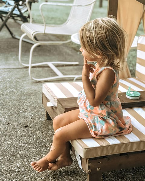 A cute little blonder girl, sitting on a sun bed, with a pondering expression.