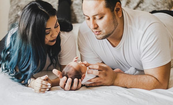 A mother and father lying on a bed, admiring their newborn baby, as the father holds her in his hands.