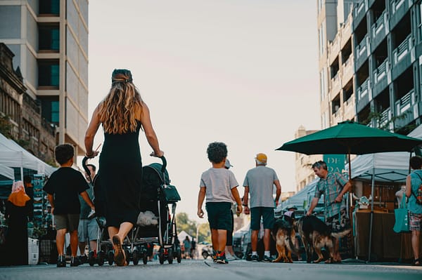 A mother pushing a double stroller through a busy outdoor marketplace on a warm afternoon.
