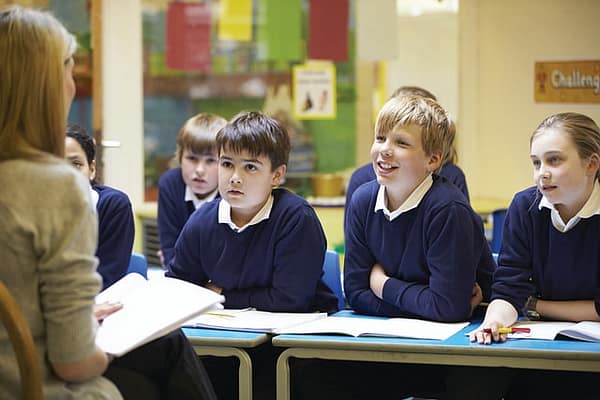 MIddle school children in matching blue school uniforms, intently listening to a teacher as she reads from a book.