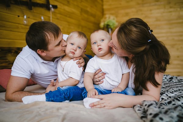 A father and mother lying on the bed with their twins boys, giving them kisses and cuddles, before their twin feeding.