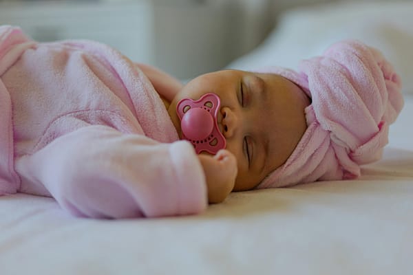 A beautiful sleeping baby girl wearing a pink robe, pink hair towel, and sucking a pink pacifier, exausted after feeding.