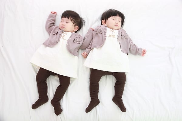 Gorgeous sleeping twins, lying side by side on a bed with white sheets, exausted after twin feeding.
