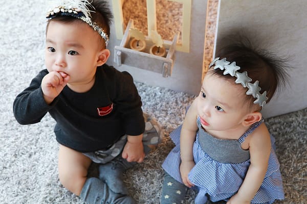 Twins babies, sitting on the floor, looking up expectantly, waiting for feeding wearing cute headbands.
