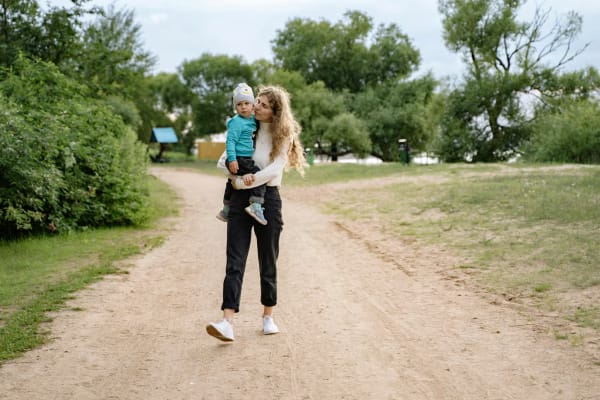 A young mother walking down a country lane, carrying her toddler in her arms, on a sunny day.