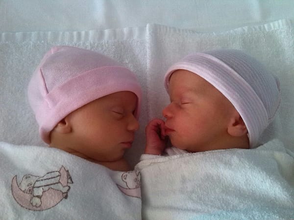 Newborn twins, peacefully lying asleep, facing each other, wearing pink beanies.