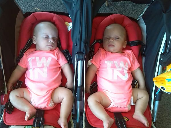 Sleeping twins baby girls in matching pink jumpsuits, asleep in a double stroller.