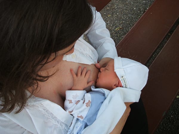 A mother breastfeeding her baby son, sitting on a park bench, outside in the cool air.