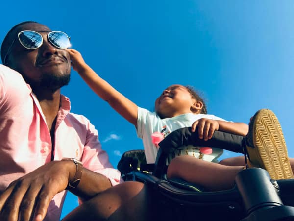 An adult wearing sunglasses and a child in a stroller are outdoors. The child is reaching toward the adult's face against a clear blue sky.