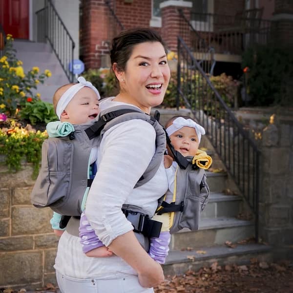 A happy mother, with smiling twin babies, carrying them in a Twingo Carrier, walking down a picturesque street.