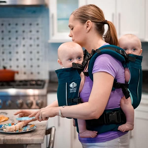 A mother holds her twins in a Twingo Carrier while cutting up a banana in the kitchen