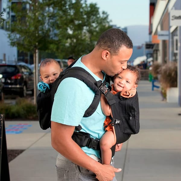 A man tenderly kisses his baby's forehead, holding his twins in a baby carrier, on a quiet street.