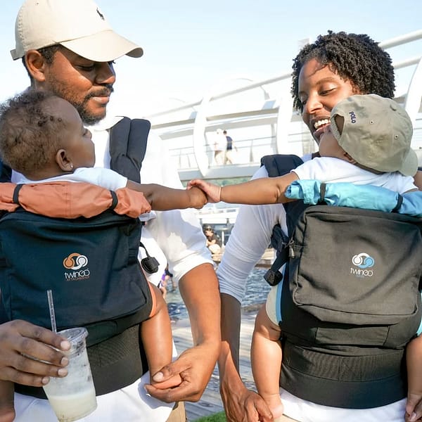 Smiling parents, outside on a sunny day, carrying their twin babies as they explore the surroundings.
