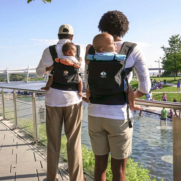 Two people outside on a sunny day, by the lake, carrying their babies on their backs in baby carriers.