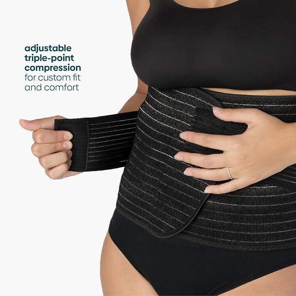 A woman in a black bra and panties, wrapping her torso in a Postpartum Belly Wrap.