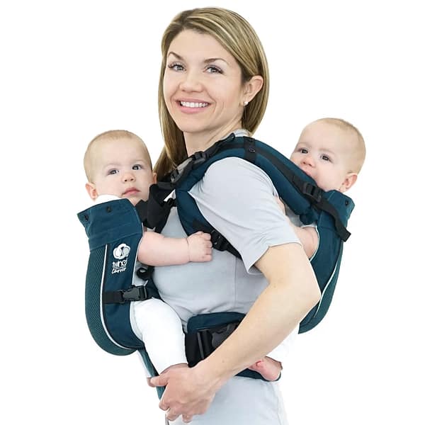 A smiling woman modelling a twin baby carrier which is holding her twin infants.