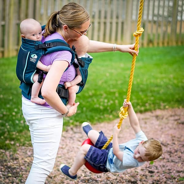 Woman wearing a Twingo carrier which is holding her twin babies, while she swings her older child on a swing in a playground.