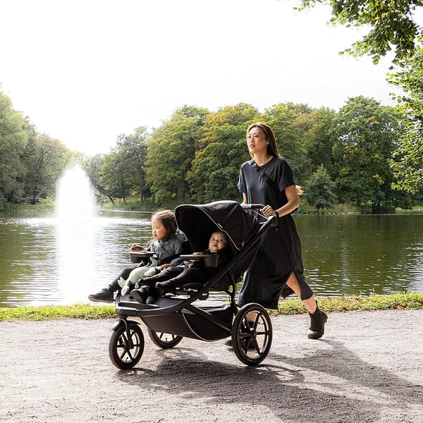 A woman happily pushing a baby in a double stroller through a sunny park.